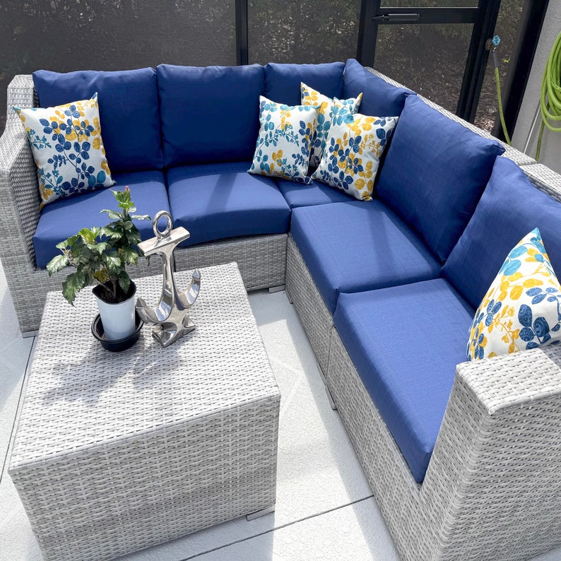May include: A light gray wicker sectional sofa with solid navy blue cushions and several throw pillows featuring a yellow and blue leaf pattern. A square wicker coffee table sits in front, holding a small potted green plant and a silver anchor.
