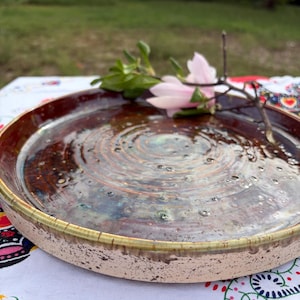 May include: A round ceramic plate with a glossy, multi-toned brown and blue glaze featuring concentric circular ripples on the interior surface. The exterior rim has a speckled cream finish with a thin, dark green edge along the top border.