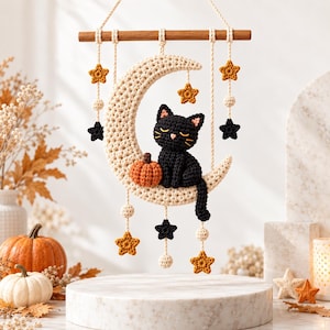 Puede incluir: Un tapiz de ganchillo hecho a mano que presenta una luna creciente de color crema, un gato negro con los ojos cerrados y una pequeña calabaza naranja. La pieza cuelga de un listón de madera mediante cuerdas adornadas con estrellas de ganchillo.