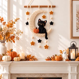May include: A crochet wall hanging featuring a cream-coloured crescent moon with a black cat and an orange pumpkin sitting on it. The moon is suspended from a wooden dowel by ropes adorned with small cream, orange, and black crocheted stars.