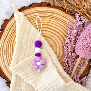 May include: A cream-coloured muslin cloth featuring a hanging accessory made of a white cord, a purple spherical bead, a white spherical bead, a smaller purple bead, and a light purple flower-shaped bead with a white centre.