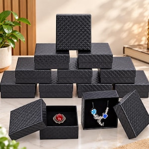 May include: A collection of square black jewelry boxes with a textured diamond pattern on the lids. One open box displays a red oval brooch, while another open box contains a silver-toned necklace with blue and silver charms inside.