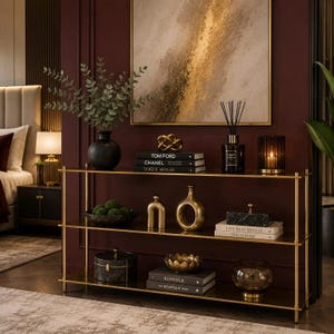 May include: A three-tier console table with a gold metal frame and dark glass shelves. The shelves are decorated with various items including books titled Tom Ford, Chanel, Vogue, Live Beautifully, and Kinfolk, along with vases, candles, and decor.