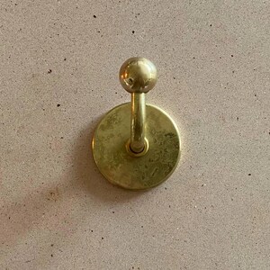 Puede incluir: Un gancho de pared circular de latón con acabado pulido y un pomo esférico en el extremo del brazo curvo. El herraje está montado sobre una placa base redonda y plana, mostrando un tono dorado metálico con sutiles variaciones en la textura.