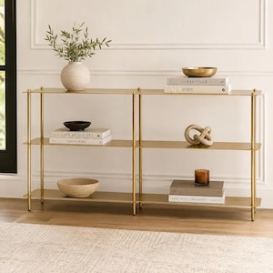 May include: A long, three-tiered console table with a gold metal frame and thin shelves. The shelves hold a white vase with greenery, a gold bowl, a decorative knot, a brown candle, and several books titled Kinfolk, AD at 100, Live Beautiful, This is Home, Aesop, and Beautifully Organized.