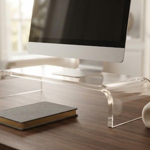 May include: A clear acrylic monitor stand with a curved design, supporting a computer screen. The stand is rectangular with rounded edges, providing a raised platform for the monitor on a wooden desk surface.