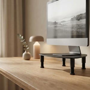 May include: A black monitor stand with a triangular platform and four adjustable cylindrical legs, supporting a computer screen displaying a black and white landscape image of hills and trees under a bright sky.