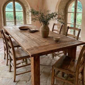Puede incluir: Una mesa de comedor rectangular de madera con textura desgastada, rodeada por cuatro sillas de madera a juego. Sobre la mesa hay un gran jarrón de cerámica lleno de ramas verdes, junto con dos cuencos de cerámica poco profundos y un cuenco pequeño.
