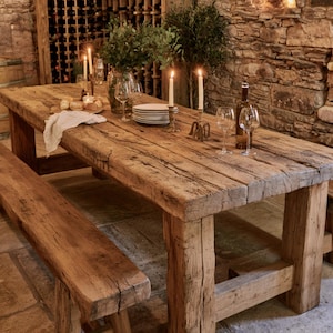 May include: A long, rectangular dining table and matching bench crafted from weathered, light brown reclaimed wood with a rough, textured surface. The table is set with white plates, clear wine glasses, two lit taper candles, and a white cloth napkin.