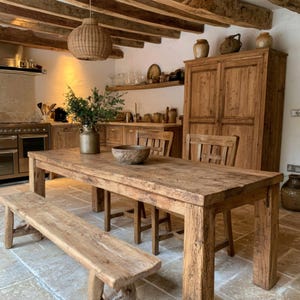 May include: A rustic dining set featuring a long, rectangular wooden table with a matching bench and two chairs. The furniture is crafted from light brown, weathered wood with visible grain. A metal pitcher with greenery sits on the table.