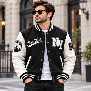 May include: A black and white varsity jacket featuring the text New York in script, the letters NY in block font, and a circular New York city skyline patch on the sleeve. The jacket has white sleeves, a black body, and striped ribbed trim.