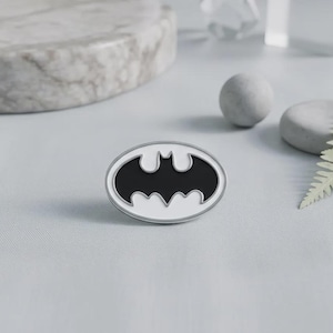 May include: An oval-shaped enamel pin featuring a black bat silhouette centred within a white background, enclosed by a thin silver-toned metal border. The design displays the iconic winged creature emblem in a clean, high-contrast graphic style.