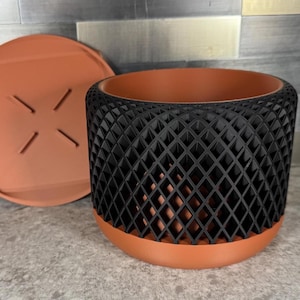 May include: A cylindrical plant pot featuring a terracotta-colored base and a removable matching saucer, paired with a black outer shell that has a geometric diamond-shaped lattice pattern. The saucer has four small raised lines in the center.