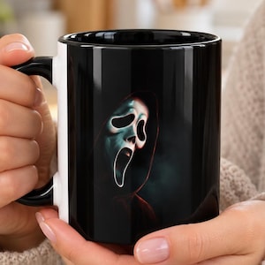 Peut inclure: Un mug en céramique noire avec une impression graphique représentant un masque de fantôme blanc allongé sous une capuche sombre. Le mug possède une anse et un intérieur noirs, mettant en valeur un design contrasté du personnage de film d'horreur.
