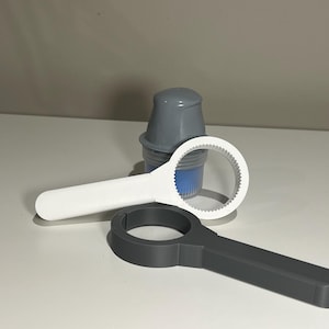 May include: A white plastic jar opener with a serrated inner ring, a dark grey plastic jar opener with a smooth inner ring, and a small grey and blue container with a screw-top lid positioned behind the two openers.