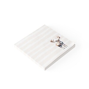May include: A square notepad featuring a light beige and white horizontal striped pattern. A watercolor illustration of a brown moose with large antlers is positioned on the right side of the top sheet of the stack of paper.