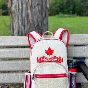 Könnte beinhalten: Ein beigefarbener Rucksack aus Segeltuch mit einem roten, gestickten Ahornblatt über einer Berg- und Waldlandschaft. Der Rucksack verfügt über eine Fronttasche mit Reißverschluss, rote Zierleisten, Seitentaschen mit Webmuster und ein SJ-Label.