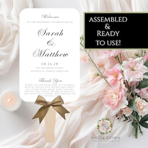 May include: A wedding fan with a gold bow and a wooden handle. The fan features the names "Sarah & Matthew", the date "08.26.28", and the location "Asheville, North Carolina". Text on the fan reads "Thank You" and "Assembled & Ready to Use!"