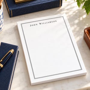 May include: A white rectangular notepad featuring a thin black double-line border and the name JOHN WILLIAMSON printed in black capital letters at the top center of the page. The notepad is composed of multiple stacked sheets of paper.