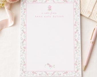 May include: A rectangular notepad featuring a delicate pink floral border and a thin pink frame. The text reads a note from ANNA KATE SUTTON in pink lettering, with a small floral motif above and below the text. A pink pen rests beside the notepad.
