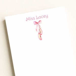 May include: A white notepad with the text "Miss Lacey" in pink script. A pink ribbon with two pink ballet shoes hanging from it is in the top right corner.