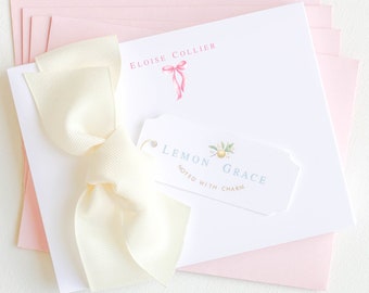 May include: A stack of light pink stationery envelopes with a white card on top. The card has a white ribbon tied in a bow and the text "ELOISE COLLIER" printed in black. A small tag with the text "LEMON GRACE NOTED WITH CHARM" is attached to the card.