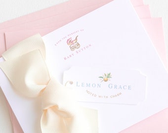May include: A stack of pink stationery envelopes with a white card on top. The card has a cream-colored bow and a small tag that says "Lemon Grace Noted with Charm". The top of the card has a small pink and white illustration of a baby carriage with the text "From the nursery of Baby Sutton".