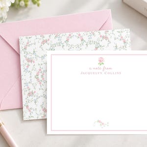 May include: A white stationery card with a pink border, a small pink rose illustration, and the text 'a note from JACQUELYN COLLINS'. It is paired with a matching envelope featuring a floral pattern of pink roses and blue flowers on white.