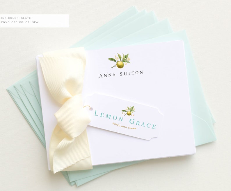 May include: A stationery set featuring a white notepad with the name "Anna Sutton" and a lemon illustration. A matching tag reads "Lemon Grace" with the words "Noted with Charm." The set is tied with a cream ribbon and includes pale green envelopes.