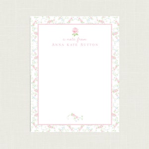 May include: A white card with a pink floral border. The card has a pink rose in the top corner and the text "a note from ANNA KATE SUTTON" in the center.