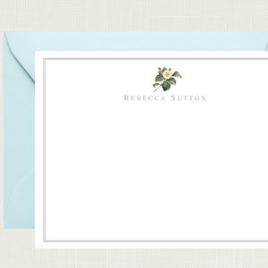 May include: A white card with a black border and a small white flower with green leaves in the top center. The text "Rebecca Sutton" is centered below the flower.