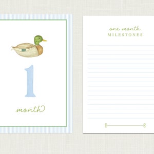 May include: A blue and white striped card with a green duck illustration and the number one in blue. The card says "one month" and has a blank space for writing. The card is next to a white card with blue lines and the text "one month milestones".