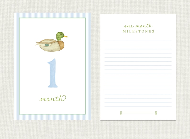May include: A blue and white striped card with a green duck illustration and the number one in blue. The card says "one month" and has a blank space for writing. The card is next to a white card with blue lines and the text "one month milestones".
