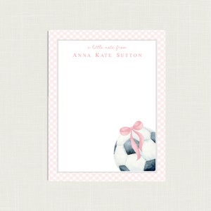 May include: A personalized notepad with a pink gingham border and a soccer ball illustration. The text "a little note from" and the name "ANNA KATE SUTTON" are printed at the top. A pink bow adorns the soccer ball.