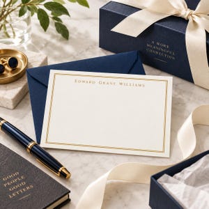 May include: A cream-colored stationery card with a gold border and the name Edward Grant Williams printed in gold. The card rests on a navy blue envelope next to a navy blue gift box with a cream ribbon and a navy blue fountain pen.