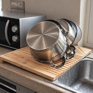 Peut inclure: Un égouttoir à vaisselle rectangulaire en bois avec des rainures parallèles, placé au-dessus d'un évier de cuisine. Plusieurs casseroles en acier inoxydable à poignées noires sont empilées et sèchent sur la planche, montrant des bases métalliques.