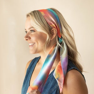 May include: A long, rectangular hair scarf featuring a vibrant, multi-coloured gradient pattern with blended hues of orange, pink, purple, and teal. The smooth, satin-like fabric is tied around the head, with two long ends hanging down the side.