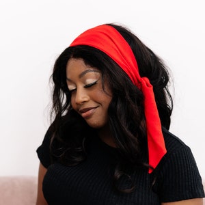 May include: A woman with long, dark curly hair wears a red headband tied in a bow at the back of her neck. She is wearing a black, ribbed, short-sleeved shirt.