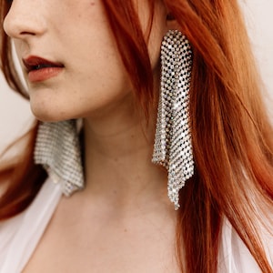 May include: A close-up of a person wearing long, red hair and large, silver, dangling earrings. The earrings are made of a chain-like material with small, sparkling rhinestones.