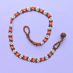 May include: A beaded necklace with a floral design. The necklace features small, round beads in white, orange, and brown, arranged in a daisy-like pattern. The necklace has a loop and a beaded ball closure. The background is a solid light purple.