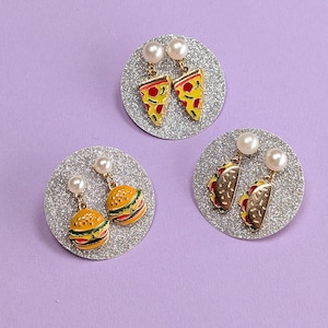 May include: Three pairs of novelty earrings on silver glitter circles. One pair features pizza slices, another hamburgers, and the last tacos. Each earring has a pearl stud. The background is a solid lavender color.