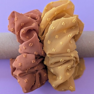 Puede incluir: Dos coleteros en tonos marrón y amarillo. Los coleteros están hechos de una tela texturizada con pequeños puntos en relieve. Los coleteros se muestran sobre un fondo morado.