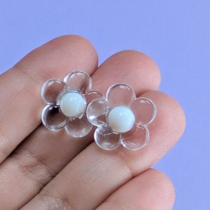 May include: Two clear flower-shaped earrings with white centers. The earrings have a delicate, transparent appearance, with each petal clearly defined. The background is a solid light purple color.