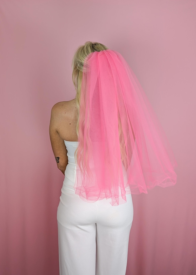 May include: A bright pink tulle veil, attached to the hair, cascades down the back. The veil is the focal point against a pink backdrop. The person is wearing a white strapless top and white pants.