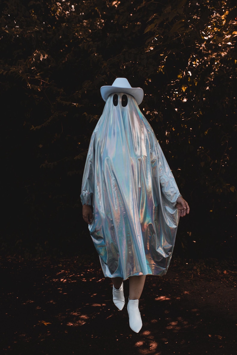 May include: A person wearing a white cowboy hat and a silver iridescent ghost costume, standing in a dark woodland setting.