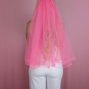 May include: A bright pink tulle veil, likely for a wedding or bachelorette party, is draped over someone's head and shoulders. The veil is a vibrant, eye-catching color, and the person is wearing a white top and pants. The background is a solid pink.