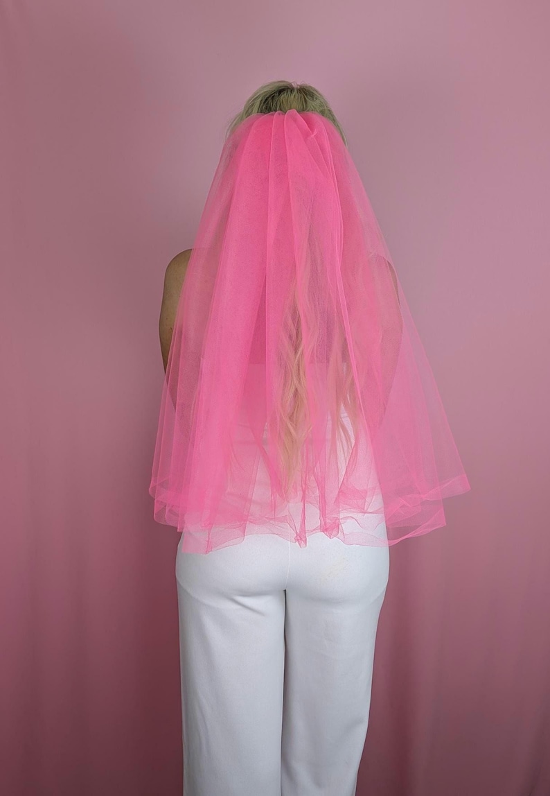 May include: A bright pink tulle veil, likely for a wedding or bachelorette party, is draped over someone's head and shoulders. The veil is a vibrant, eye-catching color, and the person is wearing a white top and pants. The background is a solid pink.