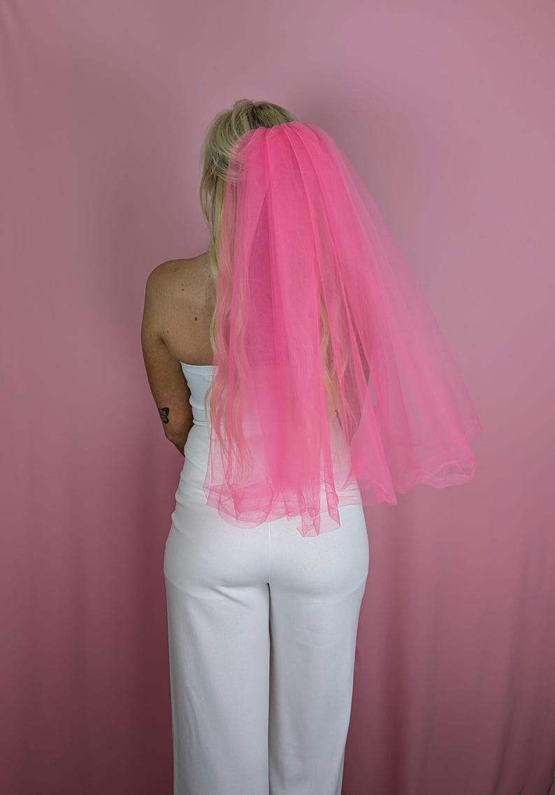 May include: A bright pink tulle veil, attached to the hair, cascades down the back. The veil is paired with a white jumpsuit against a pink backdrop. The image focuses on the veil's texture and color.