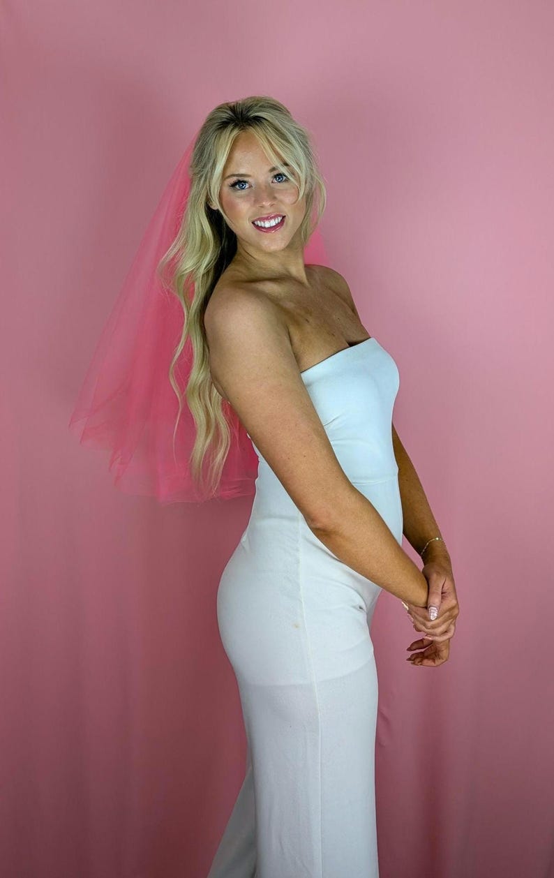 May include: A woman wearing a white strapless jumpsuit and a pink veil. The veil is a soft pink color and is attached to the woman's hair. The jumpsuit is form-fitting and has a simple, elegant design.