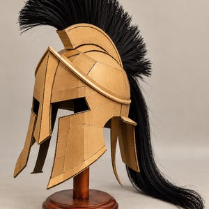 May include: A Corinthian-style helmet constructed from tan corrugated cardboard, featuring a prominent crest adorned with long, flowing black synthetic hair that extends down the back. The helmet is mounted on a circular wooden display stand.
